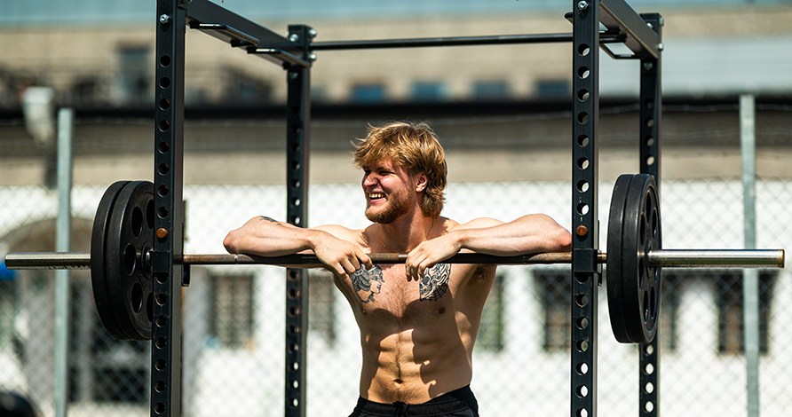 En leende man str och lutar armarna mot en skivstng i ett utomhusgym.